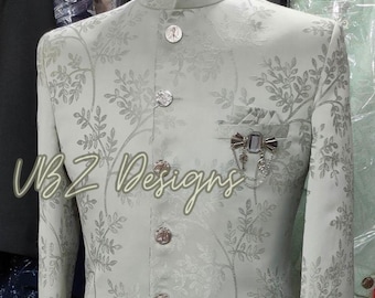 Puede incluir: Chaqueta de traje verde claro con un sutil estampado floral y botones decorativos. La chaqueta tiene cuello mandarín y un bolsillo con un alfiler decorativo. El texto "VBZ Designs" es visible en la parte delantera.