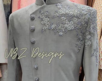 Puede incluir: Chaqueta de traje gris claro con intrincado bordado floral en el hombro y el pecho. La chaqueta presenta un cuello mandarín y botones decorativos. El texto "VBZ Designs" está escrito en cursiva en la parte inferior de la chaqueta.