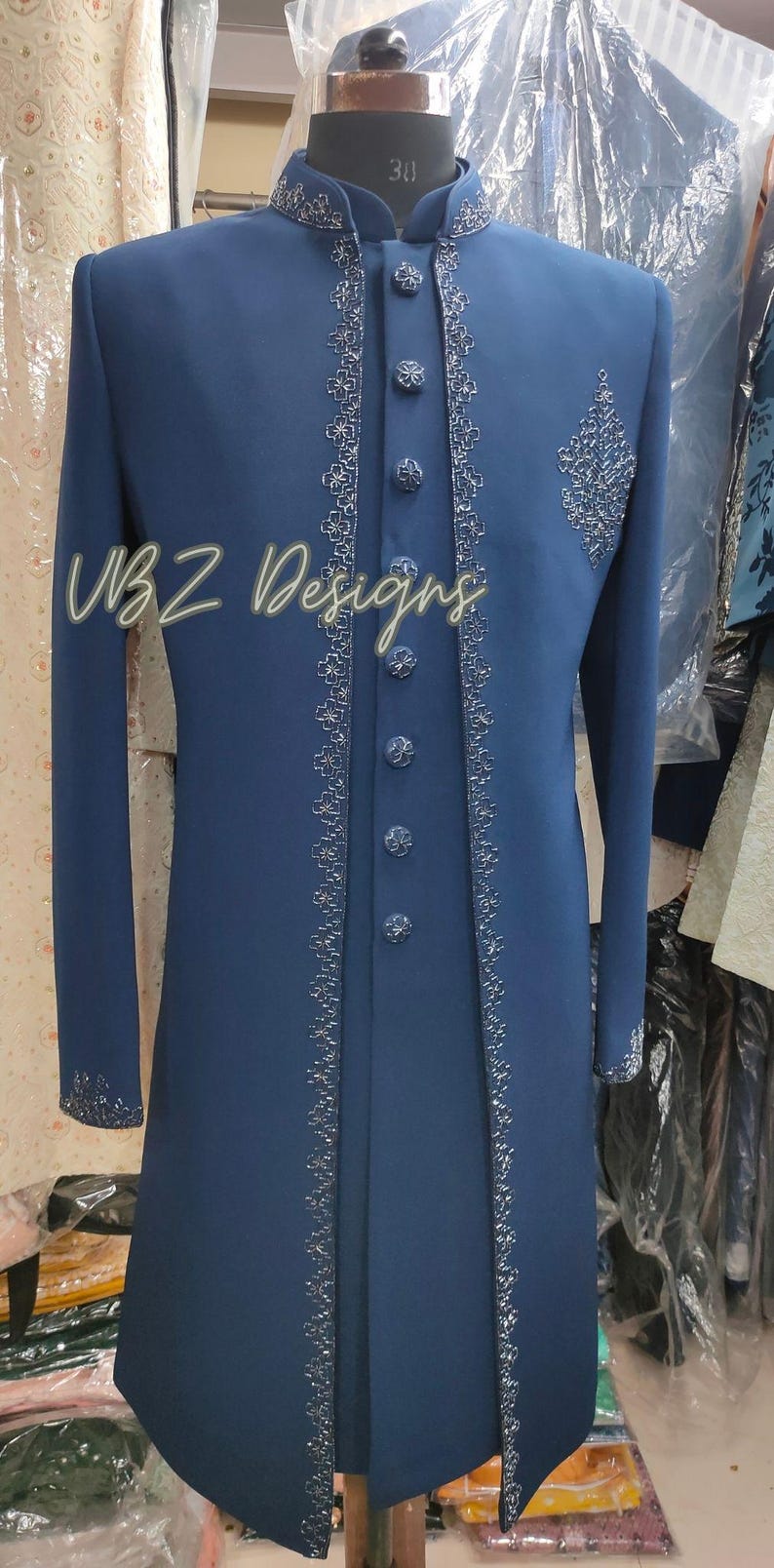 Puede incluir: Una chaqueta formal larga de color azul marino con intrincados bordados plateados en los bordes y en el pecho. La chaqueta tiene cuello mandar&iacute;n y botones decorativos. El texto "VBZ Designs" es visible.