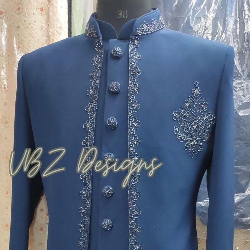 Puede incluir: Chaqueta de traje azul marino con intrincados bordados plateados alrededor del cuello, en la parte delantera y en el pecho. La chaqueta tiene botones decorativos y el texto "WBZ Designs" en cursiva.