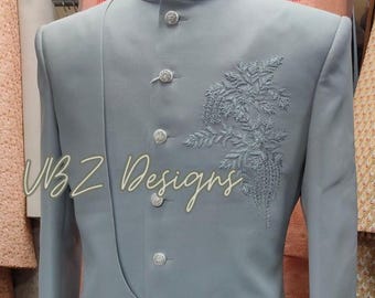 Puede incluir: Chaqueta formal gris claro con cuello mandarín y botones plateados decorativos. La chaqueta presenta un intrincado bordado floral en la parte delantera y el texto "VBZ Designs".