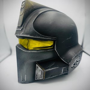 May include: A black and gray helmet with yellow visor lenses. The helmet has a weathered appearance with silver accents and a skull emblem on the top. The design is reminiscent of science fiction or futuristic armor.