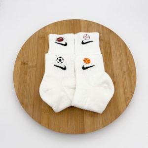 Könnte beinhalten: Vier Paar weiße Sportsocken mit Sportmotiven. Jedes Paar zeigt einen schwarzen Nike-Swoosh und einen anderen Sportball: Football, Baseball, Fußball und Basketball. Die Socken liegen auf einem Holzbrett.