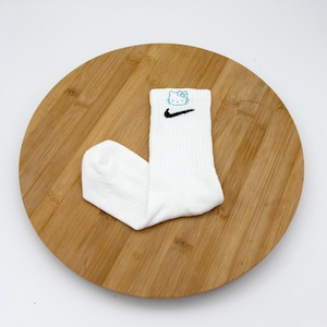 Pu&ograve; includere: Calzini sportivi bianchi con uno swoosh Nike nero e un motivo Hello Kitty blu. I calzini sono a coste e sono esposti su una superficie di legno rotonda.