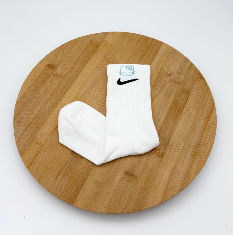 Pu&ograve; includere: Calzini sportivi bianchi con uno swoosh Nike nero e un motivo Hello Kitty blu. I calzini sono a coste e sono esposti su una superficie di legno rotonda.