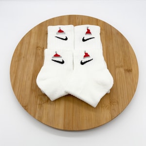 May include: A pair of white ankle socks featuring a black Nike swoosh logo embroidered on the side. Above each swoosh is a small, detailed embroidery of a red and white Santa hat. The socks are folded and presented in a clean, simple arrangement.