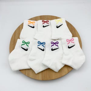 以下が含まれることがあります： カラフルなリボンが付いた白いアンクルソックス8足セット。各ソックスには黒いナイキのスウッシュロゴが付いています。