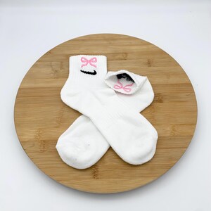 K&ouml;nnte beinhalten: Wei&szlig;e Sportsocken mit einem rosa Schleifendesign, das auf dem B&uuml;ndchen gestickt ist. Die Socken haben ein schwarzes Swoosh-Logo und werden auf einer Holzoberfl&auml;che pr&auml;sentiert. Ein l&auml;ssiges Accessoire.