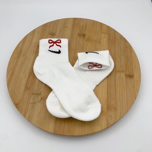 K&ouml;nnte beinhalten: Ein Paar wei&szlig;e Kn&ouml;chelsocken mit einer roten Schleife und einem schwarzen Swoosh-Logo, das auf dem B&uuml;ndchen gestickt ist. Die Socken werden auf einer runden Holzoberfl&auml;che vor wei&szlig;em Hintergrund pr&auml;sentiert. Die zweite Socke hat eine rote Schleife auf dem B&uuml;ndchen.