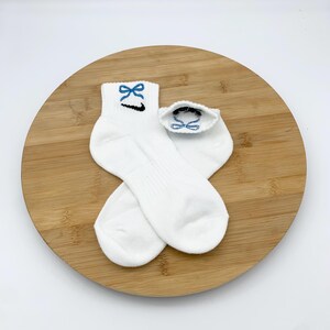 K&ouml;nnte beinhalten: Wei&szlig;e Kn&ouml;chelsocken mit einem blauen Schleifendesign, das auf dem B&uuml;ndchen gestickt ist. Das Nike Swoosh-Logo ist unterhalb der Schleife sichtbar. Die Socken werden auf einer runden Holzoberfl&auml;che pr&auml;sentiert.