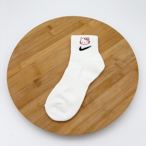 Pu&ograve; includere: Calzini alla caviglia bianchi con uno swoosh Nike nero e un motivo Hello Kitty rosso. I calzini sono realizzati in materiale morbido e sono esposti su una tavola rotonda di legno.