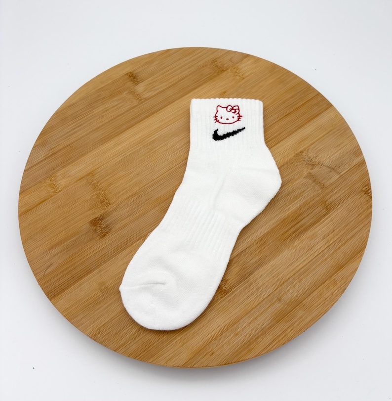 Pu&ograve; includere: Calzini alla caviglia bianchi con uno swoosh Nike nero e un motivo Hello Kitty rosso. I calzini sono realizzati in materiale morbido e sono esposti su una tavola rotonda di legno.