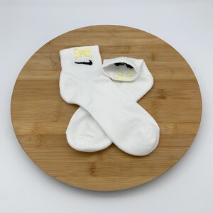 K&ouml;nnte beinhalten: Zwei wei&szlig;e Kn&ouml;chelsocken mit gelbem Schleifendesign und schwarzem Swoosh-Logo. Die Socken sind aus weichem Material und werden auf einer Holzoberfl&auml;che pr&auml;sentiert.
