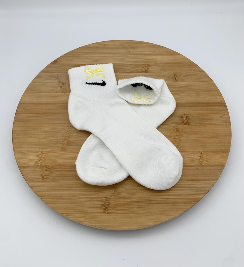 K&ouml;nnte beinhalten: Zwei wei&szlig;e Kn&ouml;chelsocken mit gelbem Schleifendesign und schwarzem Swoosh-Logo. Die Socken sind aus weichem Material und werden auf einer Holzoberfl&auml;che pr&auml;sentiert.