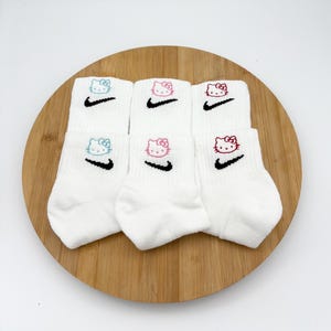 Pu&ograve; includere: Sei paia di calzini alla caviglia bianchi con facce di Hello Kitty ricamate e swoosh Nike neri. Le facce di Hello Kitty sono blu, rosa e rosse. I calzini sono disposti su una tavola rotonda di legno.