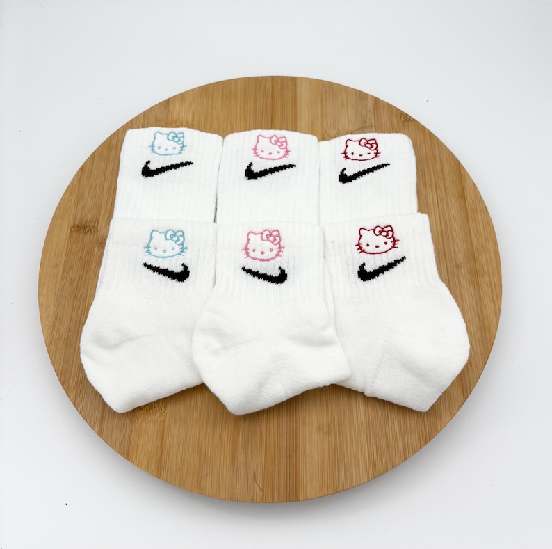 Pu&ograve; includere: Sei paia di calzini alla caviglia bianchi con facce di Hello Kitty ricamate e swoosh Nike neri. Le facce di Hello Kitty sono blu, rosa e rosse. I calzini sono disposti su una tavola rotonda di legno.
