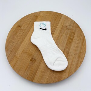 Pu&ograve; includere: Calzino alla caviglia bianco con uno swoosh Nike nero e un motivo Hello Kitty blu. Il calzino &egrave; a coste e si trova su una superficie di legno.