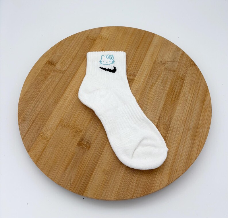 Pu&ograve; includere: Calzino alla caviglia bianco con uno swoosh Nike nero e un motivo Hello Kitty blu. Il calzino &egrave; a coste e si trova su una superficie di legno.