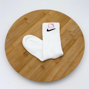 Pu&ograve; includere: Calzini sportivi bianchi con uno swoosh Nike nero e una grafica rosa di Hello Kitty. I calzini sono a coste e sembrano essere fatti di un materiale morbido. Presentati su una superficie rotonda di legno.