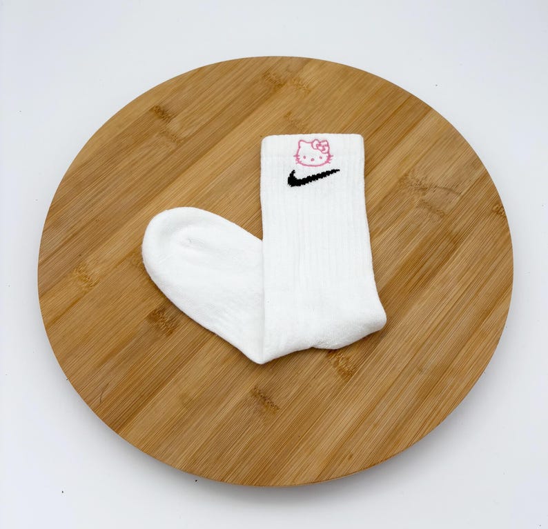Pu&ograve; includere: Calzini sportivi bianchi con uno swoosh Nike nero e una grafica rosa di Hello Kitty. I calzini sono a coste e sembrano essere fatti di un materiale morbido. Presentati su una superficie rotonda di legno.