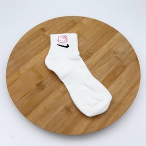 Pu&ograve; includere: Calzino alla caviglia bianco con un motivo di Hello Kitty rosa e uno swoosh Nike nero. Il calzino &egrave; realizzato in un materiale morbido ed &egrave; progettato per il comfort. Il calzino &egrave; esposto su una superficie di legno.