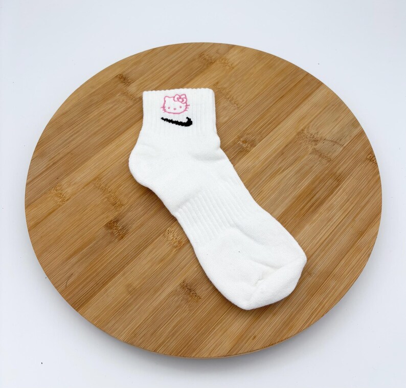 Pu&ograve; includere: Calzino alla caviglia bianco con un motivo di Hello Kitty rosa e uno swoosh Nike nero. Il calzino &egrave; realizzato in un materiale morbido ed &egrave; progettato per il comfort. Il calzino &egrave; esposto su una superficie di legno.