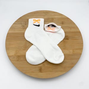 K&ouml;nnte beinhalten: Ein Paar wei&szlig;e Kn&ouml;chelsocken mit einem orangefarbenen Schleifendesign und einem schwarzen Swoosh-Logo. Die Socken werden auf einer runden Holzoberfl&auml;che vor wei&szlig;em Hintergrund pr&auml;sentiert. Die Socken sind aus weichem, geripptem Material.