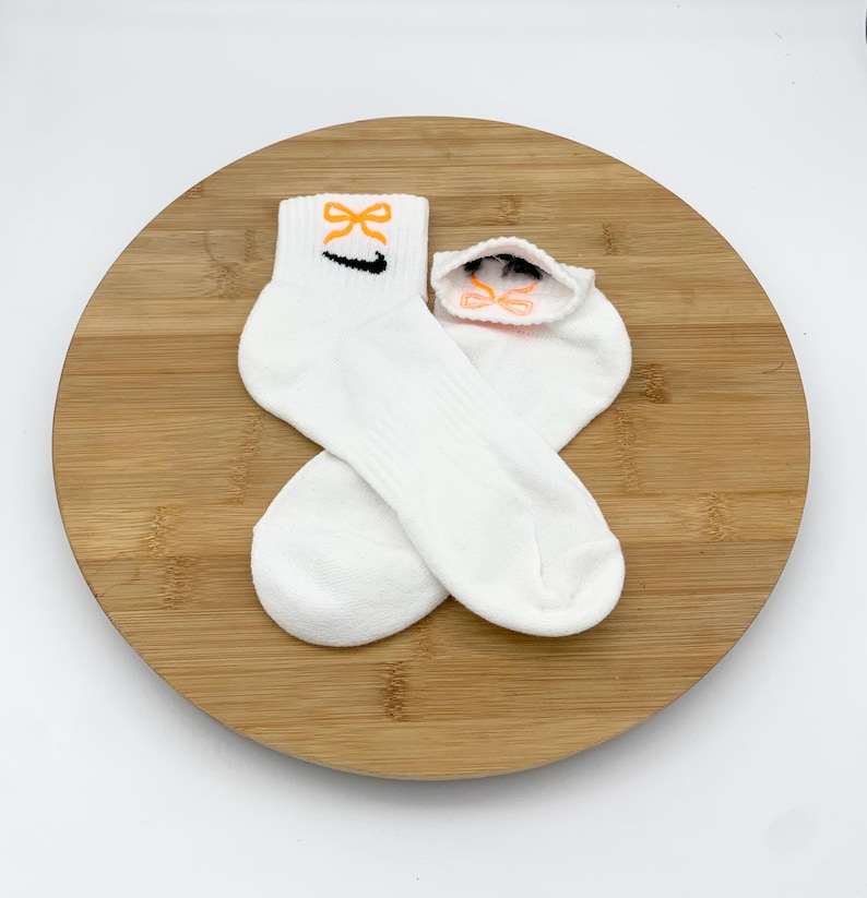 K&ouml;nnte beinhalten: Ein Paar wei&szlig;e Kn&ouml;chelsocken mit einem orangefarbenen Schleifendesign und einem schwarzen Swoosh-Logo. Die Socken werden auf einer runden Holzoberfl&auml;che vor wei&szlig;em Hintergrund pr&auml;sentiert. Die Socken sind aus weichem, geripptem Material.