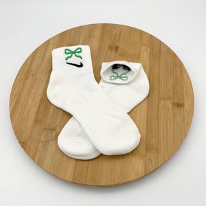 K&ouml;nnte beinhalten: Ein Paar wei&szlig;e Kn&ouml;chelsocken mit gr&uuml;ner Schleifenstickerei und einem schwarzen Swoosh-Logo. Die Socken sind aus weichem Material und werden auf einer Holzoberfl&auml;che pr&auml;sentiert.