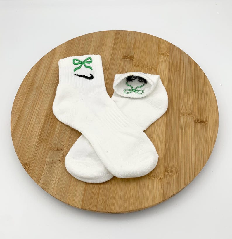 K&ouml;nnte beinhalten: Ein Paar wei&szlig;e Kn&ouml;chelsocken mit gr&uuml;ner Schleifenstickerei und einem schwarzen Swoosh-Logo. Die Socken sind aus weichem Material und werden auf einer Holzoberfl&auml;che pr&auml;sentiert.