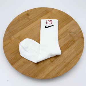Pu&ograve; includere: Calzini sportivi bianchi con uno swoosh Nike nero e una grafica di Hello Kitty rossa. I calzini sono a coste e sono su una superficie di legno.