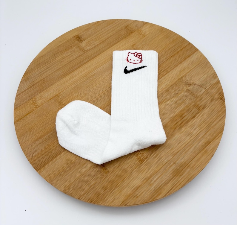 Pu&ograve; includere: Calzini sportivi bianchi con uno swoosh Nike nero e una grafica di Hello Kitty rossa. I calzini sono a coste e sono su una superficie di legno.