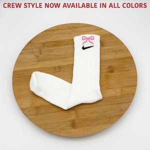 K&ouml;nnte beinhalten: Wei&szlig;e Crew-Socken mit rosa Schleife und schwarzem Nike Swoosh-Logo. Die Socken werden auf einer Holzoberfl&auml;che pr&auml;sentiert. Der Text oben lautet "CREW STYLE NOW AVAILABLE IN ALL COLORS".