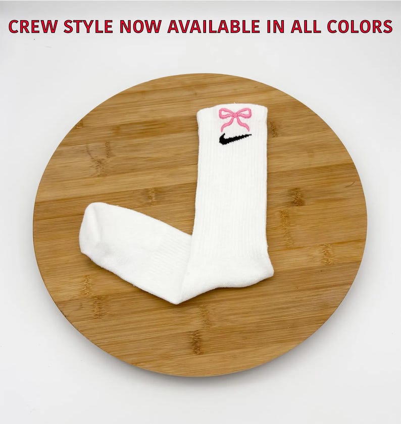 K&ouml;nnte beinhalten: Wei&szlig;e Crew-Socken mit rosa Schleife und schwarzem Nike Swoosh-Logo. Die Socken werden auf einer Holzoberfl&auml;che pr&auml;sentiert. Der Text oben lautet "CREW STYLE NOW AVAILABLE IN ALL COLORS".