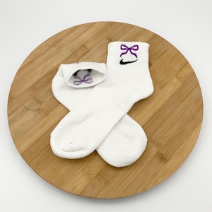 K&ouml;nnte beinhalten: Ein Paar wei&szlig;e Kn&ouml;chelsocken mit einem lila Schleifenmuster, das auf dem B&uuml;ndchen gestickt ist. Unter der Schleife befindet sich ein schwarzes Swoosh-Logo. Die Socken liegen auf einer runden Holzoberfl&auml;che.