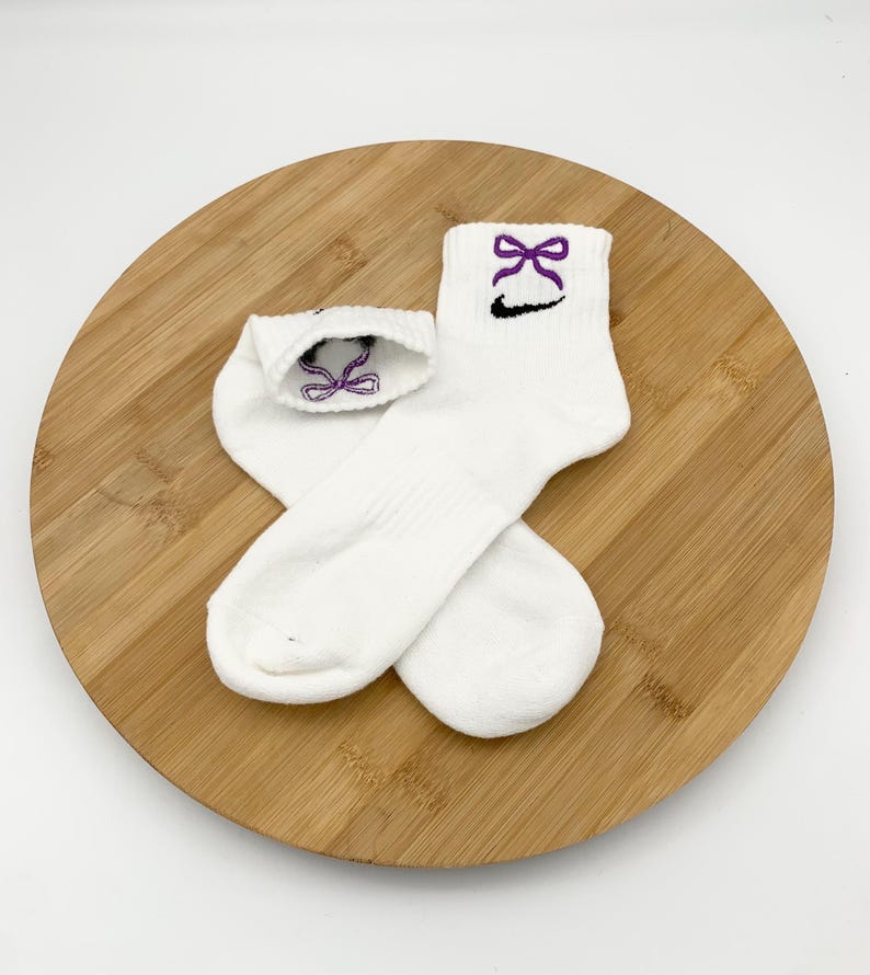 K&ouml;nnte beinhalten: Ein Paar wei&szlig;e Kn&ouml;chelsocken mit einem lila Schleifenmuster, das auf dem B&uuml;ndchen gestickt ist. Unter der Schleife befindet sich ein schwarzes Swoosh-Logo. Die Socken liegen auf einer runden Holzoberfl&auml;che.