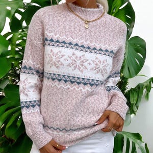 May include: A pink, long-sleeved sweater with a mock neck. The sweater features a white band with snowflake patterns and blue and pink geometric designs. The sweater is made of a soft, textured material.