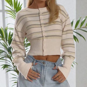 May include: Cream-coloured, cropped cardigan with horizontal stripes in light green and grey. The long-sleeved sweater has a button-up front and ribbed cuffs. The cardigan is paired with light blue denim jeans.
