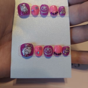 Könnte beinhalten: Ein Set mit zehn pinkfarbenen und violetten, selbstklebenden Zehennägeln mit verschiedenen Designs, darunter ein Comic-Charakter, eine Krone und Glitzer.