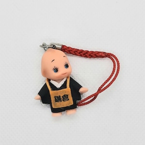 May include: A small, bald, cartoon-like figure wearing a black robe and a yellow sash with Japanese characters. The figure is attached to a red string.