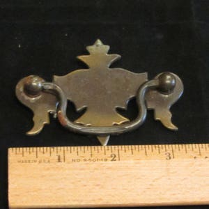 May include: A vintage brass drawer pull with a decorative design. The pull is shaped like a stylized bird with wings and a crown on its head. The pull is approximately 2 inches wide.