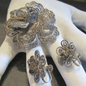 May include: A vintage silver filigree jewelry set featuring a large flower brooch and two matching flower earrings. The intricate openwork design showcases delicate floral patterns. The set is displayed on a white surface.