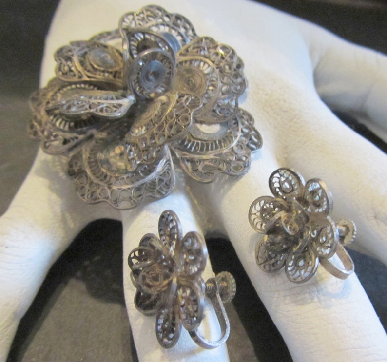 May include: A vintage silver filigree jewelry set featuring a large flower brooch and two matching flower earrings. The intricate openwork design showcases delicate floral patterns. The set is displayed on a white surface.