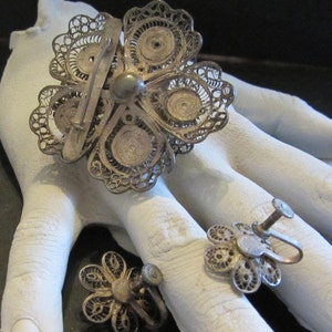 May include: A vintage silver-toned filigree flower brooch and matching screw-back earrings. The brooch is a large, openwork flower with a pin back. The earrings are smaller, also flower-shaped. The jewelry is displayed on a white surface.