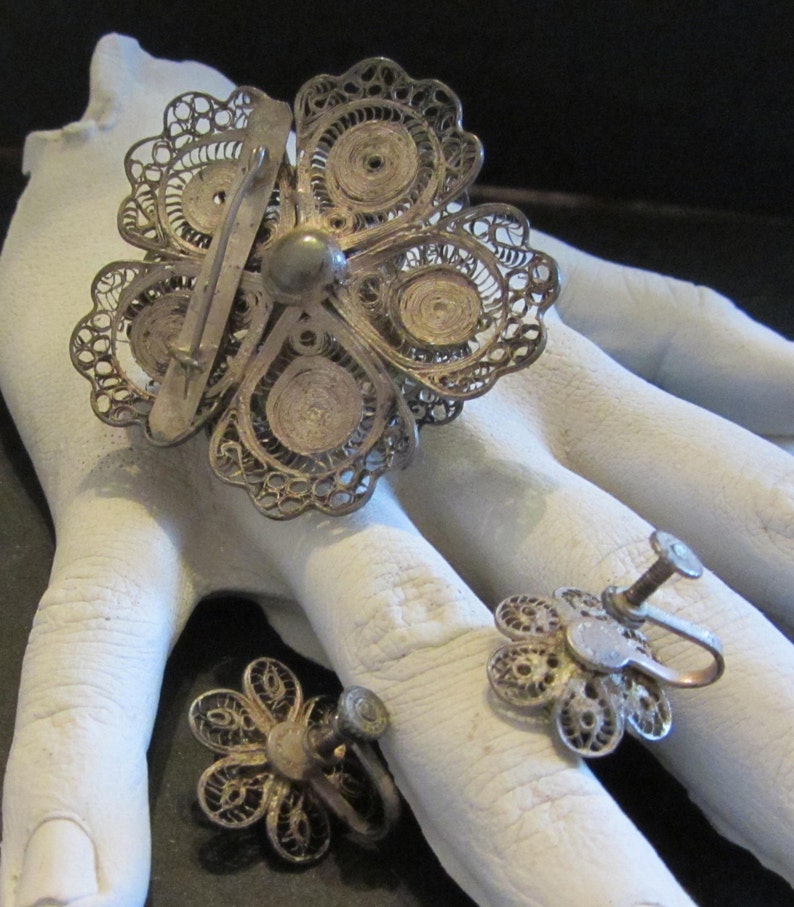 May include: A vintage silver-toned filigree flower brooch and matching screw-back earrings. The brooch is a large, openwork flower with a pin back. The earrings are smaller, also flower-shaped. The jewelry is displayed on a white surface.