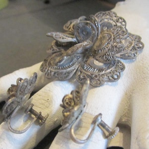 May include: A vintage silver filigree jewelry set featuring a large flower brooch and two matching screw-back earrings. The intricate openwork design showcases delicate floral patterns. The set is likely made of sterling silver.