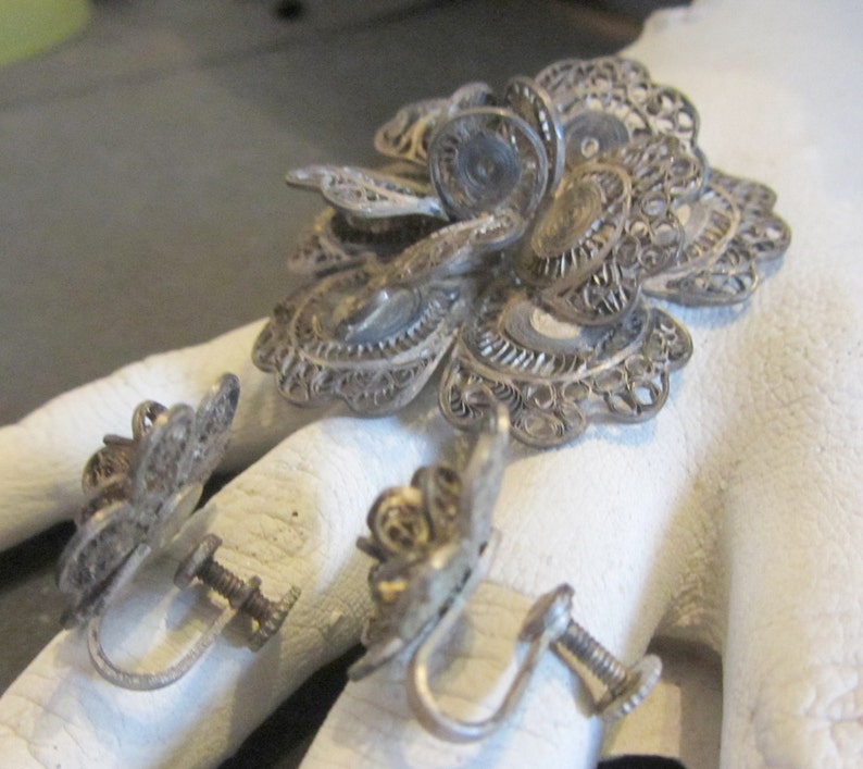 May include: A vintage silver filigree jewelry set featuring a large flower brooch and two matching screw-back earrings. The intricate openwork design showcases delicate floral patterns. The set is likely made of sterling silver.