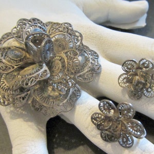 May include: A silver-toned filigree jewelry set featuring a large flower brooch and two matching flower rings. The brooch has an intricate, openwork design. The rings have a similar floral pattern. The jewelry is displayed on a white surface.