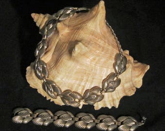 May include: A silver tone necklace and bracelet set with a leaf design. The necklace is displayed on a seashell.