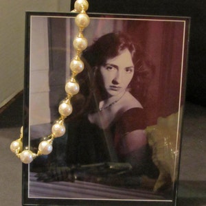 May include: A black framed photo of a woman with dark hair wearing a dark dress and a pearl necklace. The photo is set on a table with a pearl necklace draped over the frame.